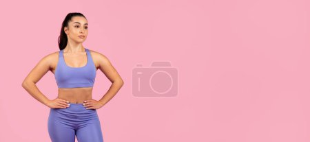 Téléchargez les photos : Une jeune femme en forme portant un soutien-gorge de sport et des leggings se tient avec confiance sur un fond rose, les mains sur les hanches - en image libre de droit