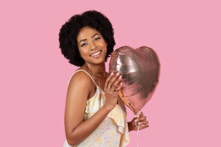 Afroamerikanerin in leichtem Sommerkleid umarmt einen herzförmigen Luftballon mit zartem Gesichtsausdruck
