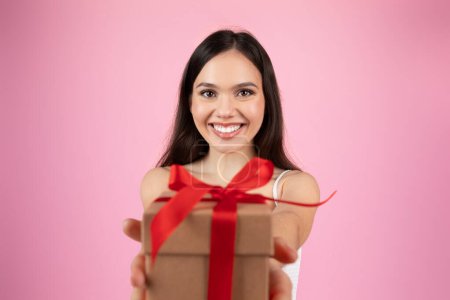 Eine entzückende junge Frau hält der Kamera eine braune Geschenkschachtel mit rotem Band auf rosa Hintergrund entgegen.