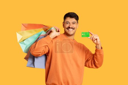 Smiling man carrying colorful shopping bags and showing a credit card, on a plain yellow background