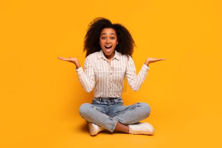 Téléchargez les photos : Joyeux jeune afro-américaine avec les mains en l'air dans la surprise, assis sur un fond de studio jaune - en image libre de droit