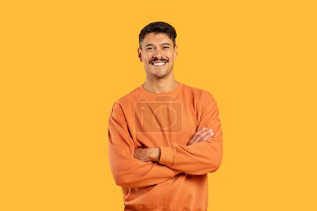 A cheerful man stands with arms confidently crossed, wearing a casual orange sweatshirt, against a solid yellow background