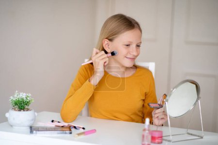 Lächelndes junges Mädchen, das mit einem Schminkpinsel Rouge aufträgt, glückliche Teenager, die sich schminken, während sie zu Hause am Tisch mit Spiegel und Kosmetik sitzen
