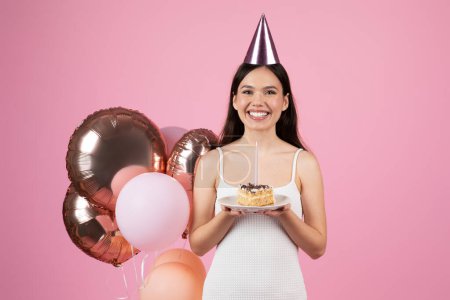 Happy young woman in a party hat holds balloons and cake, celebrating joyously on a pink background