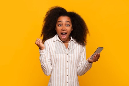 Smiling young black lady thrilled with what she sees on her phone screen against yellow backdrop