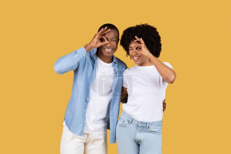 Ein fröhliches afroamerikanisches Paar, das mit den Händen vor gelbem Hintergrund spielerische Augenbewegungen macht