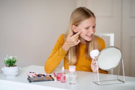 Cheerful teen girl closely examines her face in a mirror and applying moisturising face cream, female teenager focusing intently on her features, copy space