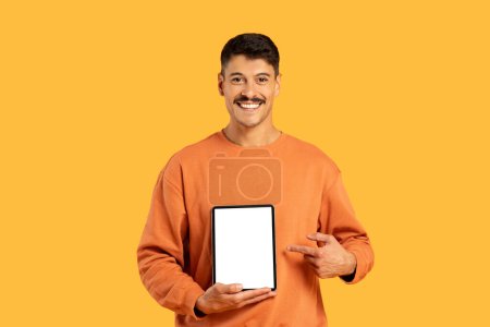 Homme joyeux présentant une tablette avec un écran blanc, vêtu d'une tenue décontractée, sur un fond jaune uniforme
