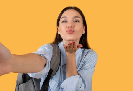 Téléchargez les photos : Une jeune femme en tenue décontractée souffle un baiser ludique, exprimant affection ou salutation sur fond jaune - en image libre de droit