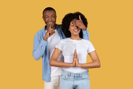 Afroamerikanischer Mann bedeckt eine Frauenauge, als sie einen hoffnungsvollen Wunsch äußert, beide stehen vor gelbem Hintergrund