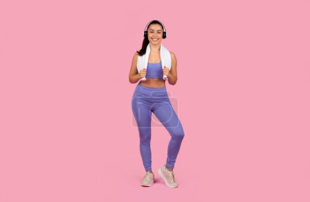A cheerful woman in sportswear with headphones poses on a pink background, showcasing an active, healthy lifestyle