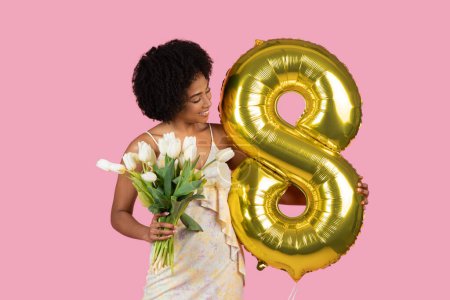A joyful young African American woman celebrates with a gold helium balloon shaped as number 8 and a bouquet of white tulips, indicating a festive occasion or achievement