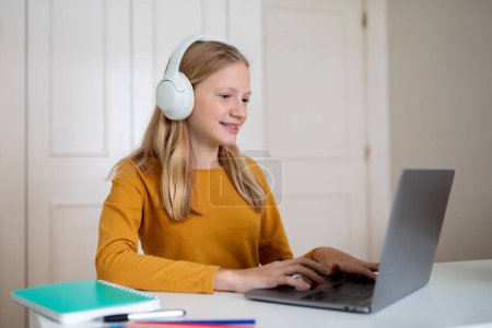 Téléchargez les photos : Jeune adolescente assise à un bureau, concentrée sur un écran d'ordinateur portable, portant un casque, étude adolescente sur ordinateur, faire des devoirs - en image libre de droit