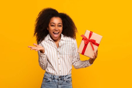 Young joyful African American woman presenting a wrapped gift box with red ribbon against a vibrant yellow background