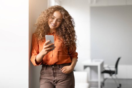 Téléchargez les photos : Une jeune femme aux cheveux bouclés utilisant son téléphone et souriant, debout dans un intérieur à la lumière naturelle - en image libre de droit