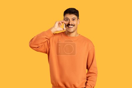 Téléchargez les photos : Joyeux jeune homme en pull orange debout sur un fond jaune, avoir une conversation téléphonique - en image libre de droit