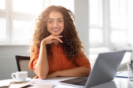 Eine selbstbewusste junge Frau mit lockigem Haar lächelt in die Kamera, während sie in einem hellen Büro an ihrem Laptop arbeitet