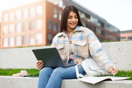 Une jeune fille concentrée dans une veste décontractée utilise attentivement son ordinateur portable assis sur un banc extérieur