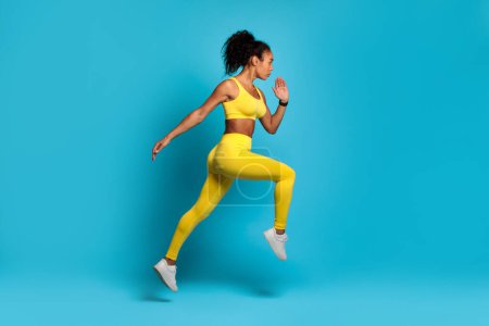 Energetic young African American woman in bright yellow sportswear captured mid-stride against a blue background