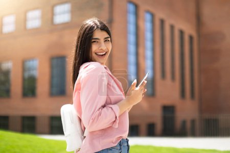 Téléchargez les photos : Une jeune étudiante sourit en regardant son téléphone, portant un sac à dos sur fond de bâtiments du campus - en image libre de droit