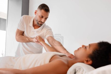 Foto de Un masajista profesional dando un relajante masaje de espalda a una mujer afroamericana cliente, envuelta en una toalla en un moderno spa o centro de bienestar - Imagen libre de derechos