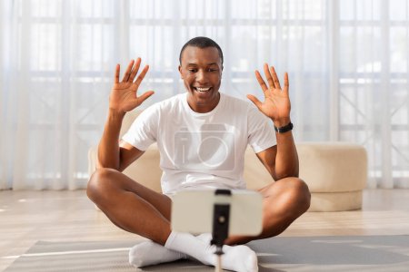 Foto de El hombre sonriente afroamericano se sienta con las piernas cruzadas en una esterilla de yoga, saludando a alguien con un gesto de manos levantadas durante una clase virtual - Imagen libre de derechos