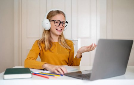 Photo for A cheerful teen girl with glasses and headphones gestures while participating in an online conversation on her laptop, happy female teenager attending virtual lesson, sitting at desk at home - Royalty Free Image