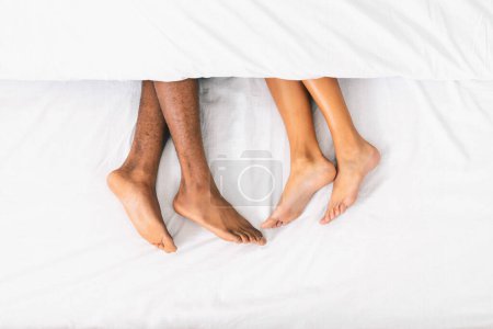 Photo for Black male and female feet lying on bed under blanket, top view with free space - Royalty Free Image