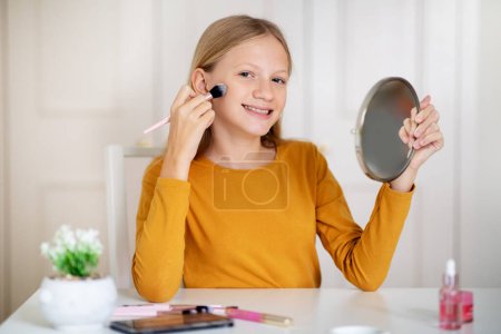 Teenager-Mädchen, die zu Hause mit einem Schminkpinsel Rouge auftragen, glückliche weibliche Teenager, die Spiegel halten und in die Kamera lächeln und Schönheitsroutine genießen