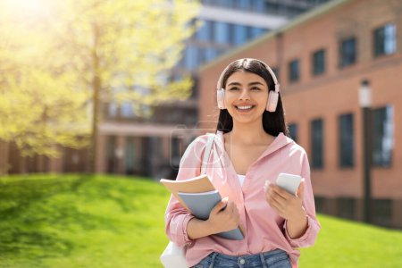 Téléchargez les photos : Une jeune étudiante portant un casque sourit vivement avec un bâtiment du campus derrière elle, tenant des livres et un téléphone - en image libre de droit