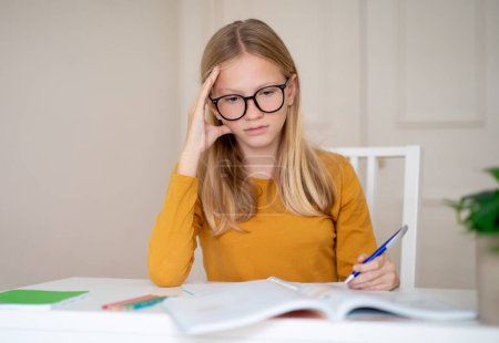 Focused young teen girl wearing glasses is studying from a book at a table, female teenager holding pen and looking to workbook, doing school homework