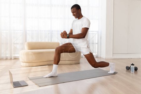 Téléchargez les photos : Un homme noir concentré maintient l'équilibre et l'équilibre en faisant une fente sur un tapis de yoga dans un espace de vie bien éclairé et minimaliste - en image libre de droit