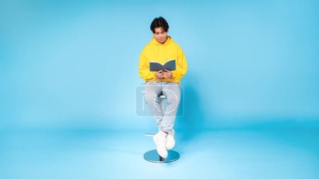 Engrossed in reading, a young asian guy sits on a stool with a book against a blue studio background