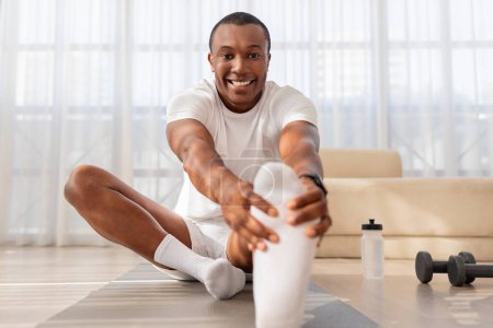 Foto de Un hombre afroamericano sonriente en ropa deportiva está estirando su pierna mientras está sentado en una esterilla de yoga en el interior, mostrando un estilo de vida saludable y una rutina de fitness en el interior. - Imagen libre de derechos