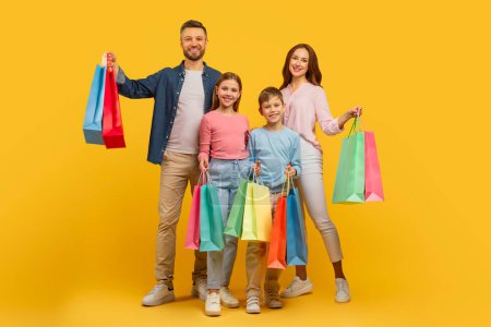 Téléchargez les photos : Père de famille mère et deux enfants profitant d'une frénésie de shopping avec des sacs colorés, sur fond jaune - en image libre de droit