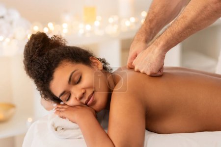 Téléchargez les photos : Cadre paisible d'une heureuse femme afro-américaine bénéficiant d'un massage des épaules apaisant au salon de spa - en image libre de droit