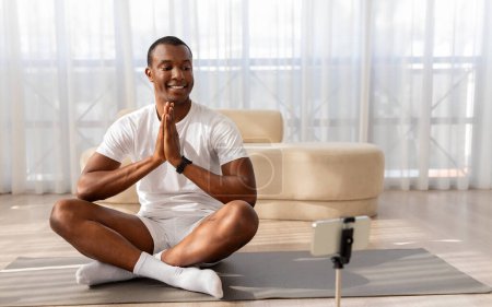 Foto de Hombre afroamericano en camiseta blanca y pantalones se sienta con las piernas cruzadas en una esterilla de yoga, grabándose con un teléfono inteligente en un trípode para la clase en línea - Imagen libre de derechos