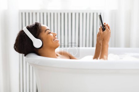 Foto de La imagen representa una relajación moderna con tecnología, mujer afroamericana en una bañera absorta en su teléfono inteligente - Imagen libre de derechos