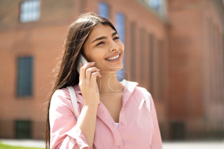 Téléchargez les photos : Une étudiante joyeuse bavarde sur un smartphone, avec un fond ensoleillé et une architecture de campus visibles - en image libre de droit