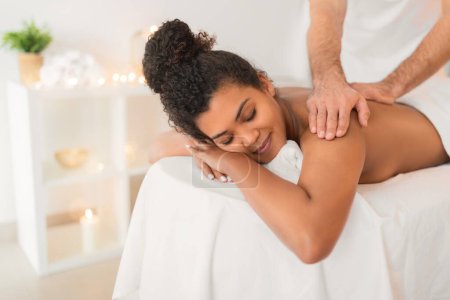 Téléchargez les photos : Une image capturant un masseur offrant un massage des épaules à une femme afro-américaine détendue allongée sur une table de spa - en image libre de droit