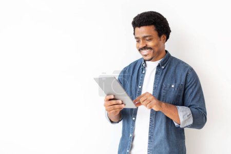 Photo for Content black man in denim shirt engaging with digital tablet, cheerful young african american male using modern gadget, standing against white backdrop, perfect for tech and lifestyle themes - Royalty Free Image