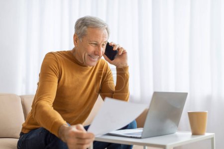 Reifer Mann im Senfpullover schaut aufmerksam auf Papiere, während er zu Hause telefoniert, mit Laptop