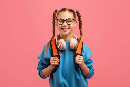 Téléchargez les photos : Enfant souriant fille mignonne avec des cheveux tressés portant des écouteurs et portant un sac à dos sur fond rose - en image libre de droit