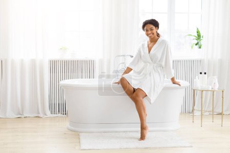 Téléchargez les photos : Dame afro-américaine sereine en peignoir pose avec une élégance décontractée sur le bord d'une baignoire élégante dans une salle de bain lumineuse - en image libre de droit