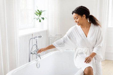 Téléchargez les photos : Femme afro-américaine dans une robe blanche luxueuse atteint pour le robinet, des éléments élégants soulignent la salle de bain moderne - en image libre de droit