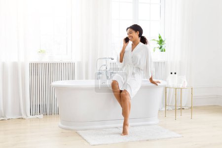 Téléchargez les photos : Joyeuse femme afro-américaine en peignoir a une conversation téléphonique éclairée dans une salle de bain nette et propre - en image libre de droit