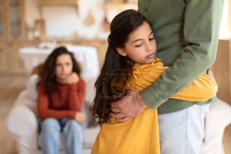Ein kleines Kind umarmt den Vater, während die Mutter im Hintergrund sitzt und zu Hause aufgebracht oder eifersüchtig wirkt