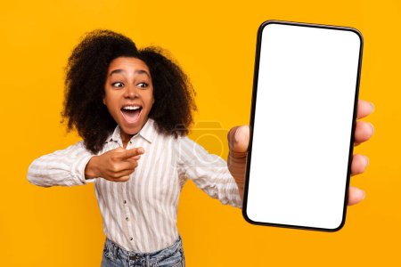 A cheerful young African American woman with curly hair pointing at a blank smartphone screen, isolated on a yellow background