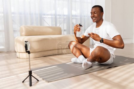 Foto de Hombre afroamericano sonríe mientras graba un video en su teléfono montado en un trípode, sentado en una esterilla de yoga en una sala de estar bien iluminada - Imagen libre de derechos