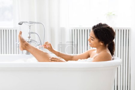African American woman enjoys a tranquil moment alone in a white bathtub, depicting self-care and relaxation in a bright bathroom, shaving legs